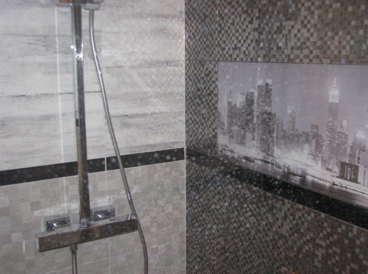Kabina prysznicowa z szaro-białymi płytkami imitującymi drewno i mozaiką z motywem panoramy Nowego Jorku, widoczna chromowana bateria.