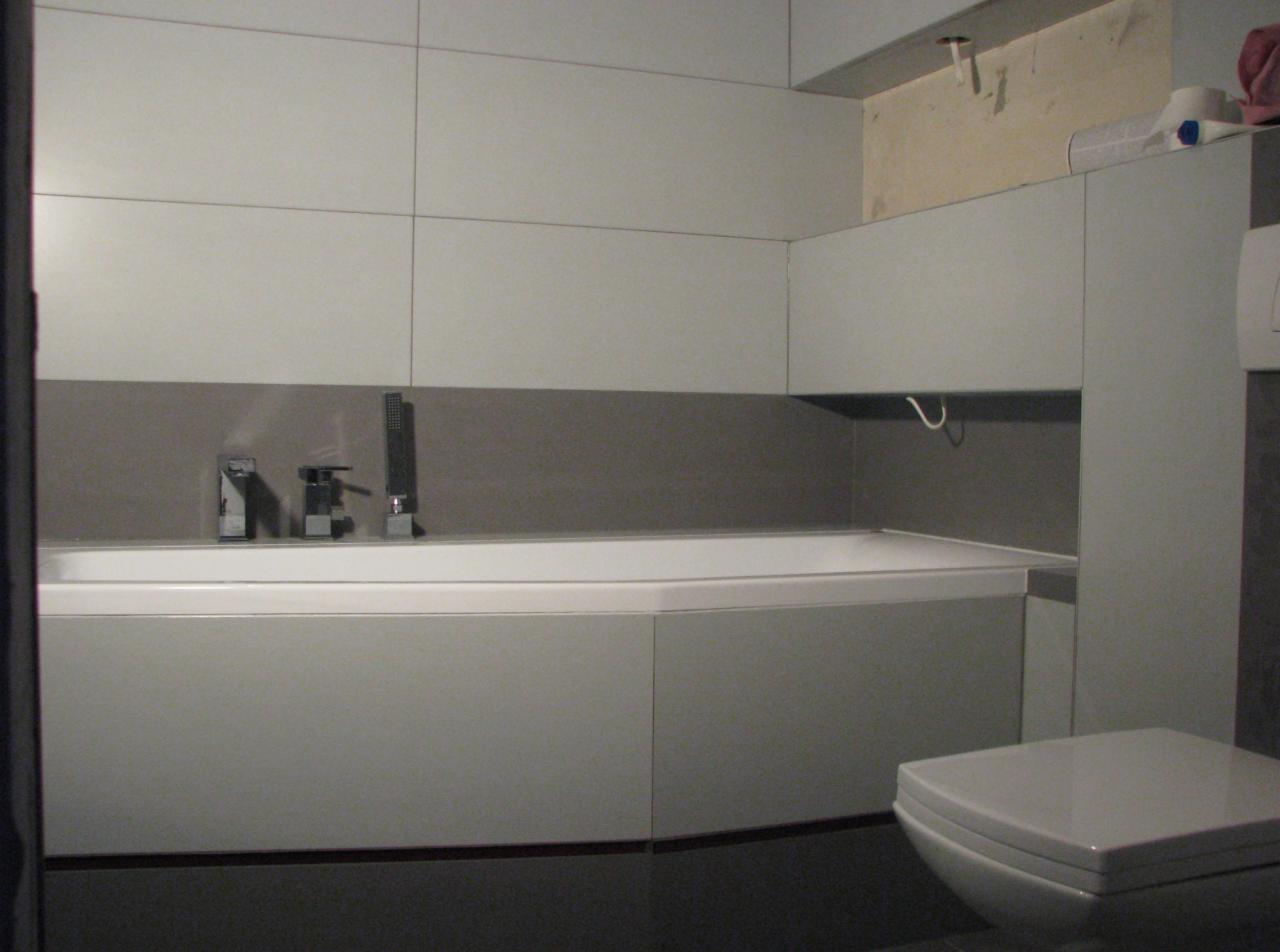 Nowoczesna łazienka w szarościach z wanną zabudowaną panelami i armaturą, widoczne płytki na ścianach i podwieszana szafka, w tle niedokończone wykończenie sufitu.