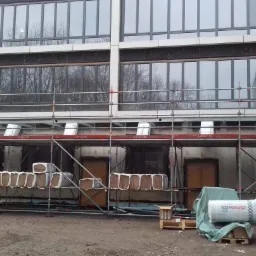 Budynek w trakcie ocieplania, widoczne rusztowania, materiał izolacyjny w rolkach i paczkach, okna z szarymi ramami, teren budowy.