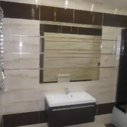 Wykończona łazienka z beżowo-brązowymi płytkami, lustrem w ciemnej ramie nad białą umywalką z szafką, grzejnikiem drabinkowym i kabiną prysznicową z ciemnymi elementami.