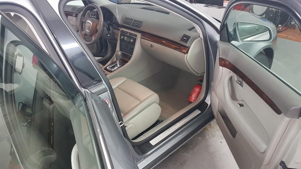 Wnętrze srebrnego Audi A4 z beżową skórzaną tapicerką, widok na deskę rozdzielczą z drewnianymi akcentami, otwarte drzwi kierowcy i pasażera, gaśnica pod siedzeniem pasażera.