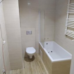 PAWEŁ RATAJCZAK - Wykończona łazienka z beżowymi płytkami imitującymi drewno na podłodze i ścianach, biała wanna z parawanem, toaleta i grzejnik drabinkowy.