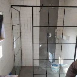 Nowoczesna kabina prysznicowa z czarnymi profilami i szklanymi ściankami w łazience wyłożonej białymi płytkami. Widoczny fragment wiadra z zaprawą na podłodze.