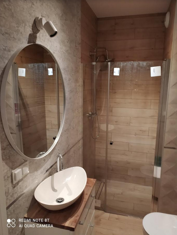 Nowoczesna łazienka z owalną umywalką nablatową, kabiną prysznicową ze szklanymi drzwiami i drewnopodobnymi płytkami na ścianach oraz podłodze.