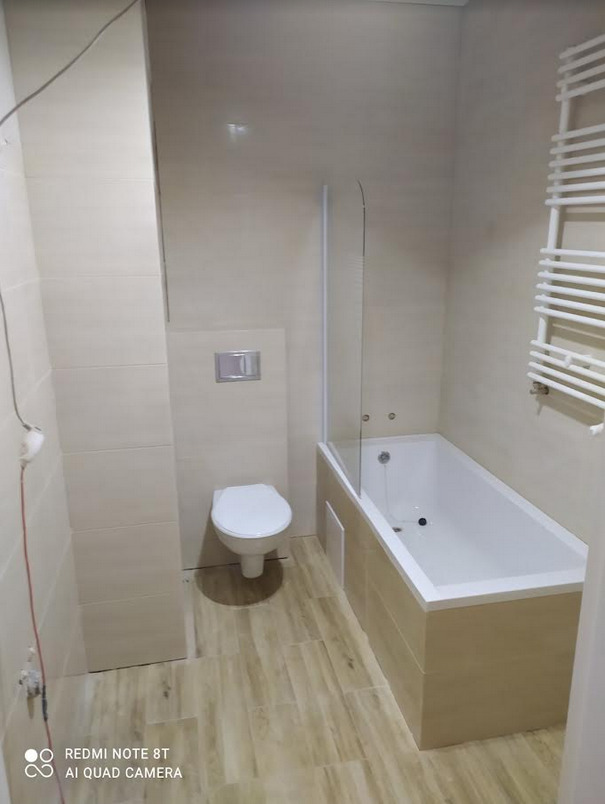 Wykończona łazienka z beżowymi płytkami imitującymi drewno na podłodze i ścianach, biała wanna z parawanem, toaleta i grzejnik drabinkowy.