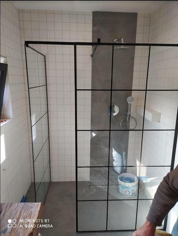 Nowoczesna kabina prysznicowa z czarnymi profilami i szklanymi ściankami w łazience wyłożonej białymi płytkami. Widoczny fragment wiadra z zaprawą na podłodze.