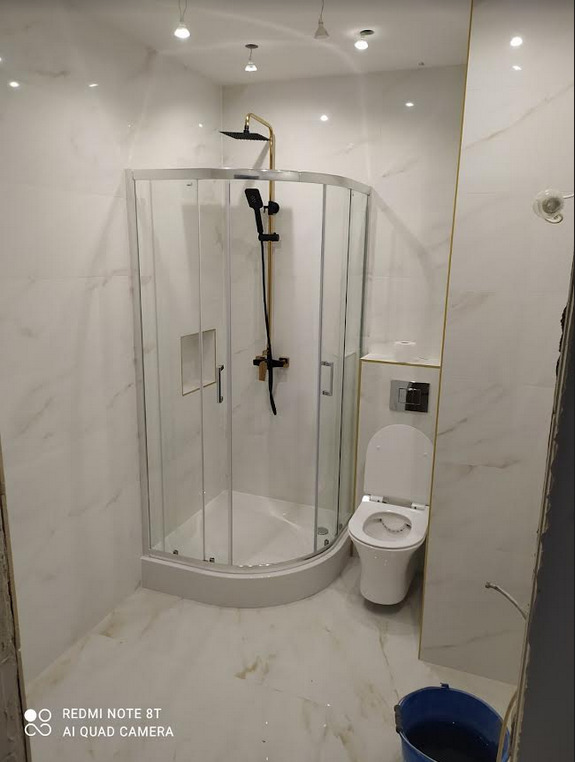 Nowoczesna łazienka z kabiną prysznicową narożną, wykończona białymi płytkami imitującymi marmur, z czarną armaturą i złotymi akcentami, widoczna toaleta i niebieskie wiadro.