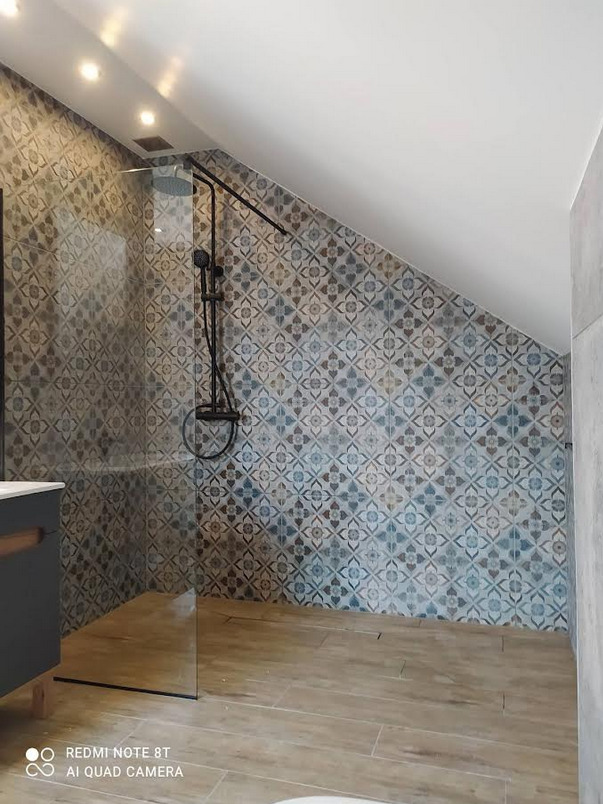 Nowoczesna łazienka na poddaszu z kabiną prysznicową typu walk-in, wykończona płytkami w stylu vintage i drewnopodobnymi panelami podłogowymi, oświetlona punktowymi lampami.