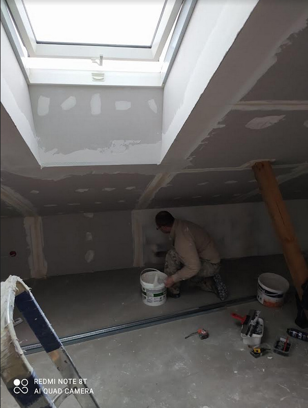 Pracownik w trakcie szpachlowania ścian poddasza z oknem dachowym, widoczne narzędzia i materiały budowlane, surowy stan wykończenia.