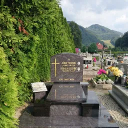 Nowoczesny, ciemny pomnik z granitu z wygrawerowanym krzyżem i zdjęciami zmarłych, na tle cmentarza z widokiem na góry.