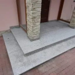 Granitowe schody wejściowe z trzema stopniami prowadzące do drzwi, z kolumną wykończoną kamieniem dekoracyjnym po lewej stronie i brukowaną ścieżką z boku.