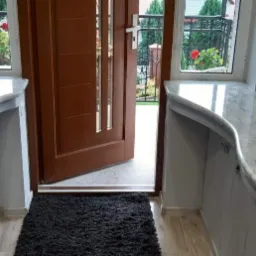 Wykończenie wnętrza wejścia z dwoma marmurowymi blatami o zaokrąglonych krawędziach po obu stronach drzwi wejściowych, widoczny pistolet do silikonu na krześle obok blatu po lewej stronie.