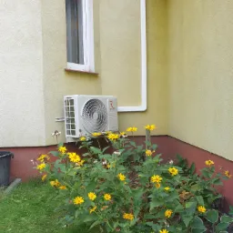 Zewnętrzna jednostka klimatyzacji Rotenso zamontowana na elewacji budynku w Żninie, otoczona kwitnącymi żółtymi kwiatami i zielenią.