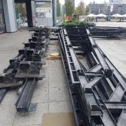 Czarne, stalowe elementy konstrukcyjne ułożone na chodniku w przestrzeni miejskiej, przygotowane do montażu, z widocznymi elementami mocującymi i śladami użytkowania.