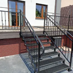 Zewnętrzne schody metalowe z czarną balustradą, prowadzące na drewniany taras przed domem z oknami z brązowymi ramami.