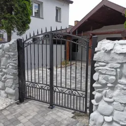 Czarna, metalowa brama wjazdowa z ozdobnymi detalami, osadzona w białym, kamiennym murze, prowadząca na posesję z domem i zadaszoną konstrukcją z drewnianymi belkami.