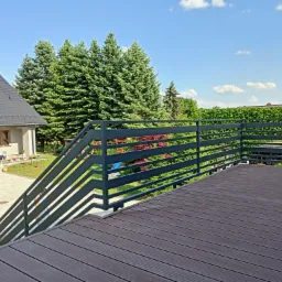 Nowoczesna, pozioma balustrada metalowa w kolorze grafitowym, zamontowana na tarasie z deski kompozytowej, z widokiem na zielony ogród i dom.