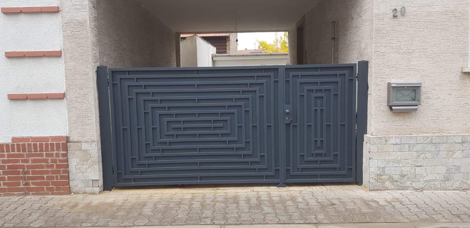 Nowoczesna, szara brama wjazdowa z geometrycznym wzorem labiryntu, zamontowana w prześwicie między budynkami z widoczną skrzynką pocztową po prawej stronie.