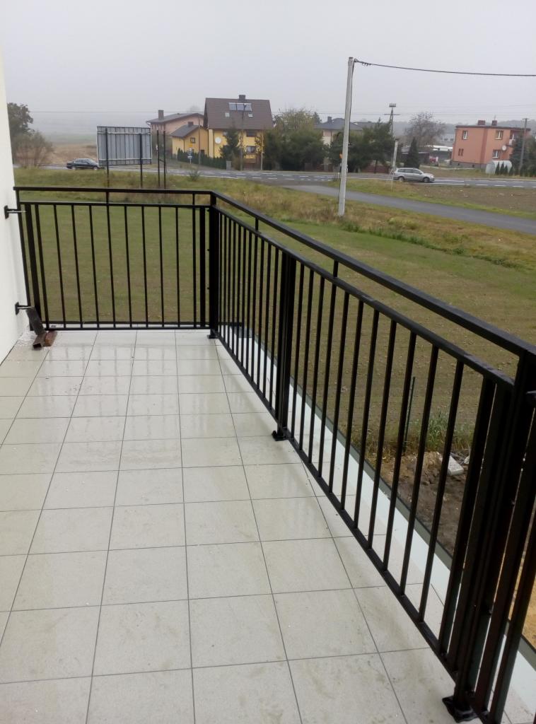 Czarna, metalowa balustrada balkonowa z pionowymi szczeblami i poziomymi poręczami, zamontowana na błyszczących, kwadratowych płytkach w Miasteczku Śląskim, z widokiem na ulicę i zabudowania w tle.
