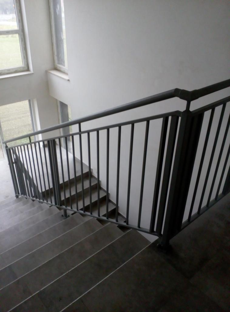Wewnętrzne schody z szarymi, betonowymi stopniami i ciemną metalową balustradą z pionowymi prętami, widok z góry w dół, minimalizm.