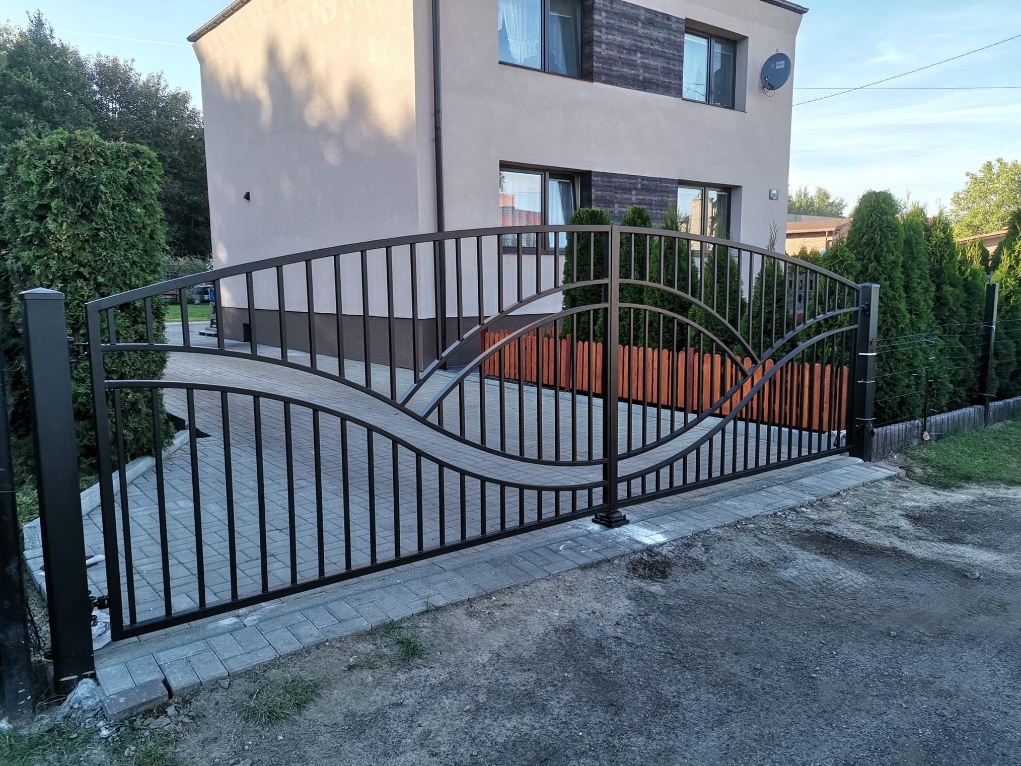 Nowoczesna brama wjazdowa z czarnego metalu o falistym wzorze, na tle domu z szarą elewacją i zielonymi tujami.