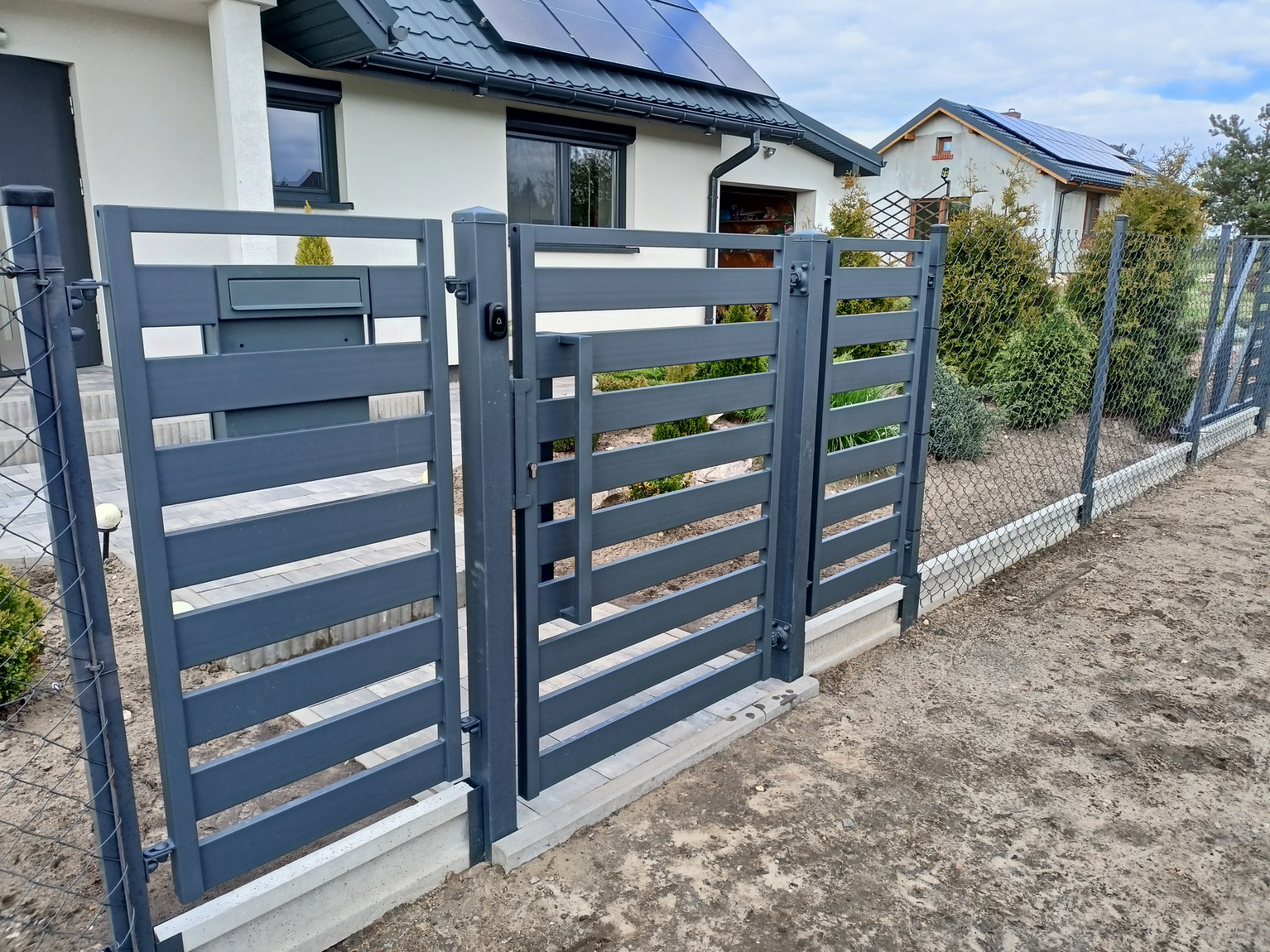 Nowoczesna brama wjazdowa z poziomych, szarych profili, zintegrowana ze skrzynką na listy, na tle domu z panelami słonecznymi.