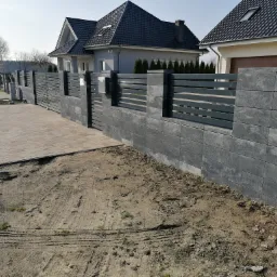 Nowoczesne ogrodzenie z szarej kostki betonowej z poziomymi przęsłami, świeżo ułożona kostka brukowa na podjeździe, widoczny fragment domu w tle, teren wokół ogrodzenia jeszcze niezagospodarowany.