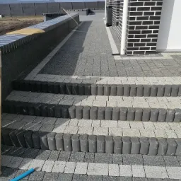 Ułożona szara kostka brukowa z obrzeżami w kształcie walców, tworząca schody i chodnik prowadzący do budynku z czarno-białą elewacją z cegły, widoczny niebieski wąż.