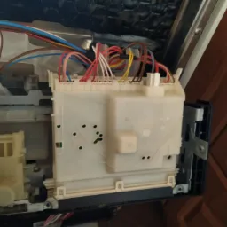 Wnętrze zmywarki z widocznym modułem sterującym i wiązką przewodów o różnych kolorach podłączonych do niego. Widoczne elementy konstrukcyjne z tworzywa sztucznego i metalu.