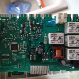 Zielona płytka drukowana z licznymi elementami elektronicznymi, w tym kondensatory, rezystory, układy scalone i przekaźniki, trzymana w dłoni, widoczne oznaczenia producenta i parametry komponentów.