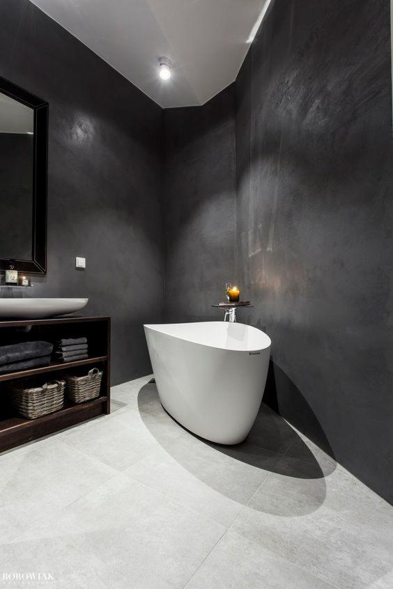 Nowoczesna łazienka z ciemnymi ścianami, wolnostojącą białą wanną, umywalką na drewnianej szafce i szarymi płytkami podłogowymi, oświetlona świecą.