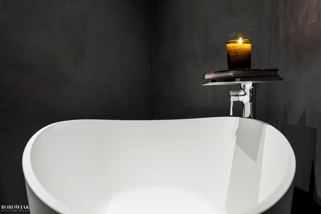 Minimalistyczna łazienka z wolnostojącą białą wanną, szarą ścianą z efektem betonu i chromowaną baterią. Na półce nad wanną leży książka i zapalona świeca w szklanym słoiku.