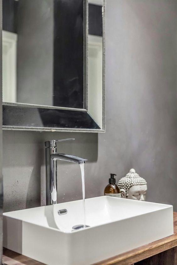 Nowoczesna łazienka z białą, prostokątną umywalką, chromowaną baterią z otwartym strumieniem wody, lustrem w srebrnej ramie i dekoracyjną figurką głowy Buddy obok dozownika mydła.