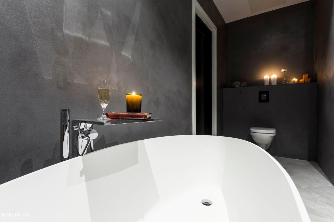 Nowoczesna łazienka z grafitowymi ścianami, wolnostojąca biała wanna, chromowana bateria, szklana półka z kieliszkiem szampana i świecą, dekoracyjne oświetlenie świecami.