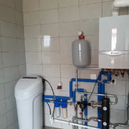 Nowoczesna kotłownia z białym kotłem gazowym Vaillant, szarym zbiornikiem wyrównawczym i filtrem wody, połączonymi rurami z niebieską izolacją na tle jasnych, kwadratowych płytek.