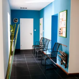 Poczekalnia w jasnych błękitnych kolorach z rzędem metalowych krzeseł, tablicą ogłoszeń i stojakiem na ulotki.