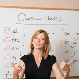 Nauczycielka angielskiego z blond włosami, w czarnej bluzce, prezentuje słówka pytające na białej tablicy, wskazując na nie ołówkiem i gestykulując dłonią z uniesionym kciukiem.