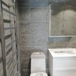 Nowoczesna łazienka z szarymi płytkami imitującymi kamień, białą szafką z umywalką, lustrzaną szafką wiszącą i chromowanym grzejnikiem drabinkowym.