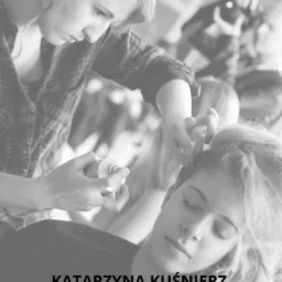 Stylistka w kraciastej koszuli, spryskuje włosy klientki siedzącej z zamkniętymi oczami, przygotowując fryzurę.