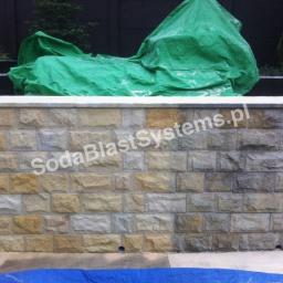 F.U.H. KRISPELL Krzysztof Szostak - Fragment kamiennego muru ogrodzeniowego, częściowo oczyszczonego z zabrudzeń, z widocznym efektem różnicy w kolorze kamienia, przykryty zieloną plandeką, z naniesionym znakiem wodnym.