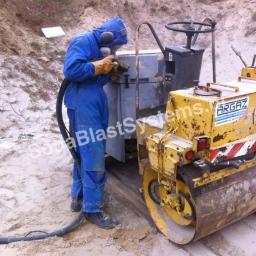 F.U.H. KRISPELL Krzysztof Szostak - Pracownik w kombinezonie ochronnym i słuchawkach, za pomocą węża z dyszą, czyści metalową obudowę maszyny budowlanej, obok stoi żółty walec drogowy.