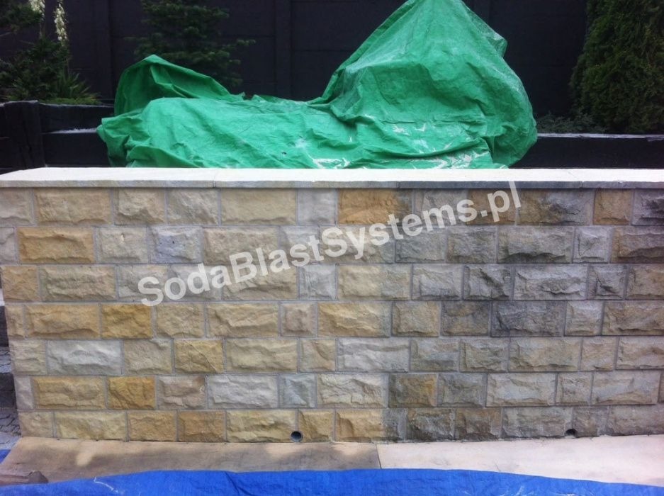 Fragment kamiennego muru ogrodzeniowego, częściowo oczyszczonego z zabrudzeń, z widocznym efektem różnicy w kolorze kamienia, przykryty zieloną plandeką, z naniesionym znakiem wodnym.