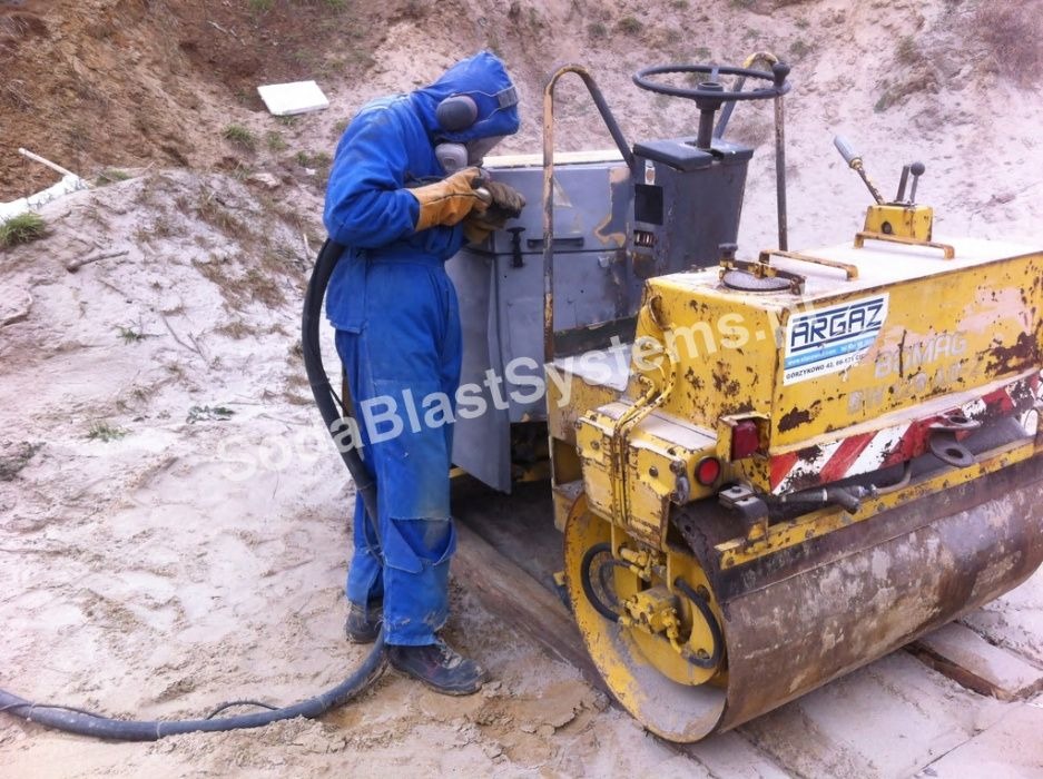 Pracownik w kombinezonie ochronnym i słuchawkach, za pomocą węża z dyszą, czyści metalową obudowę maszyny budowlanej, obok stoi żółty walec drogowy.