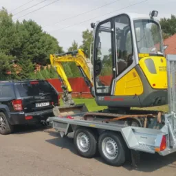 Czarny Jeep Grand Cherokee ciągnie na lawecie małą, żółto-szarą koparkę Wacker Neuson ET20.