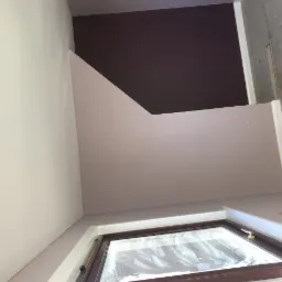 Pokój po malowaniu z oknem na poddaszu, ściany w odcieniach bieli i fioletu, widoczny fragment grzejnika i surowy beton na ścianie z prawej strony.