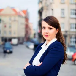 Kobieta w granatowej marynarce z założonymi rękami na tle ulicy z budynkami i samochodami, lekko pochylona w lewo.