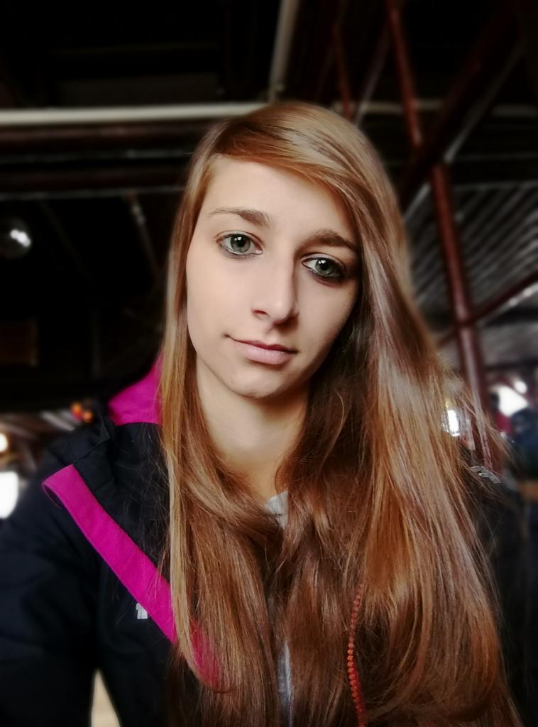 Portret młodej kobiety z długimi, kasztanowymi włosami w czarnej kurtce z różowym akcentem, na rozmytym tle z elementami metalowej konstrukcji i oświetlenia.