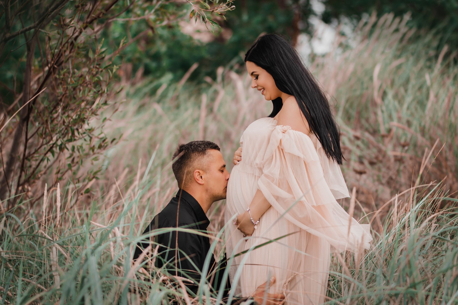 Przyszły ojciec całuje ciążowy brzuszek kobiety w zwiewnej sukni na tle traw i drzew.