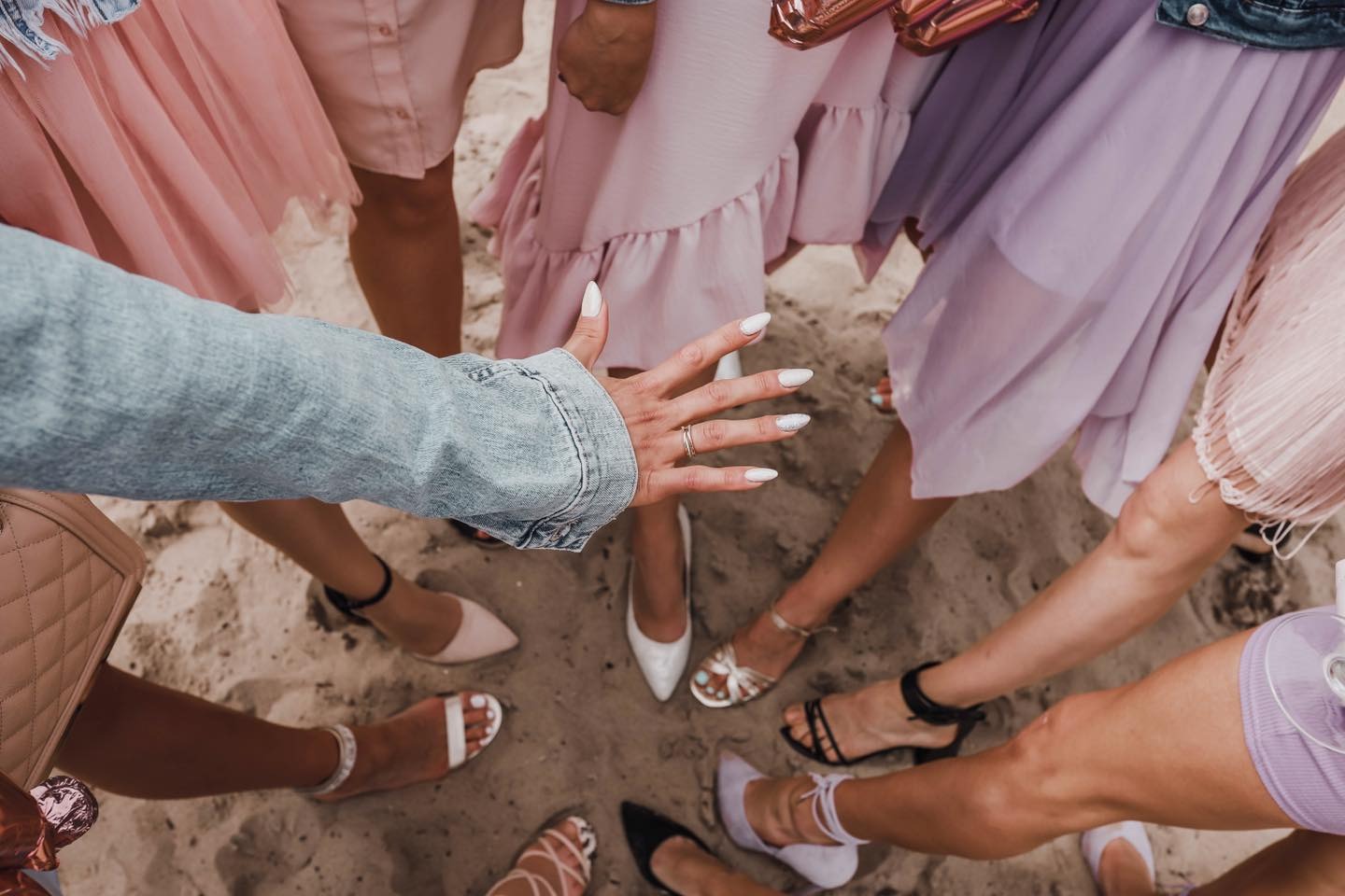 Grupa kobiet w pastelowych sukienkach i różnorodnych butach stoi na piasku, widok z góry, dłoń z pierścionkiem i pomalowanymi paznokciami w centrum kadru.