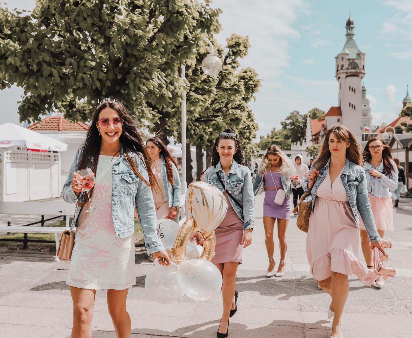 Grupa kobiet w pastelowych sukienkach i jeansowych kurtkach spaceruje po placu miejskim, trzymając balony i drinki, w tle widoczna wieża dawnego kasyna w Sopocie.